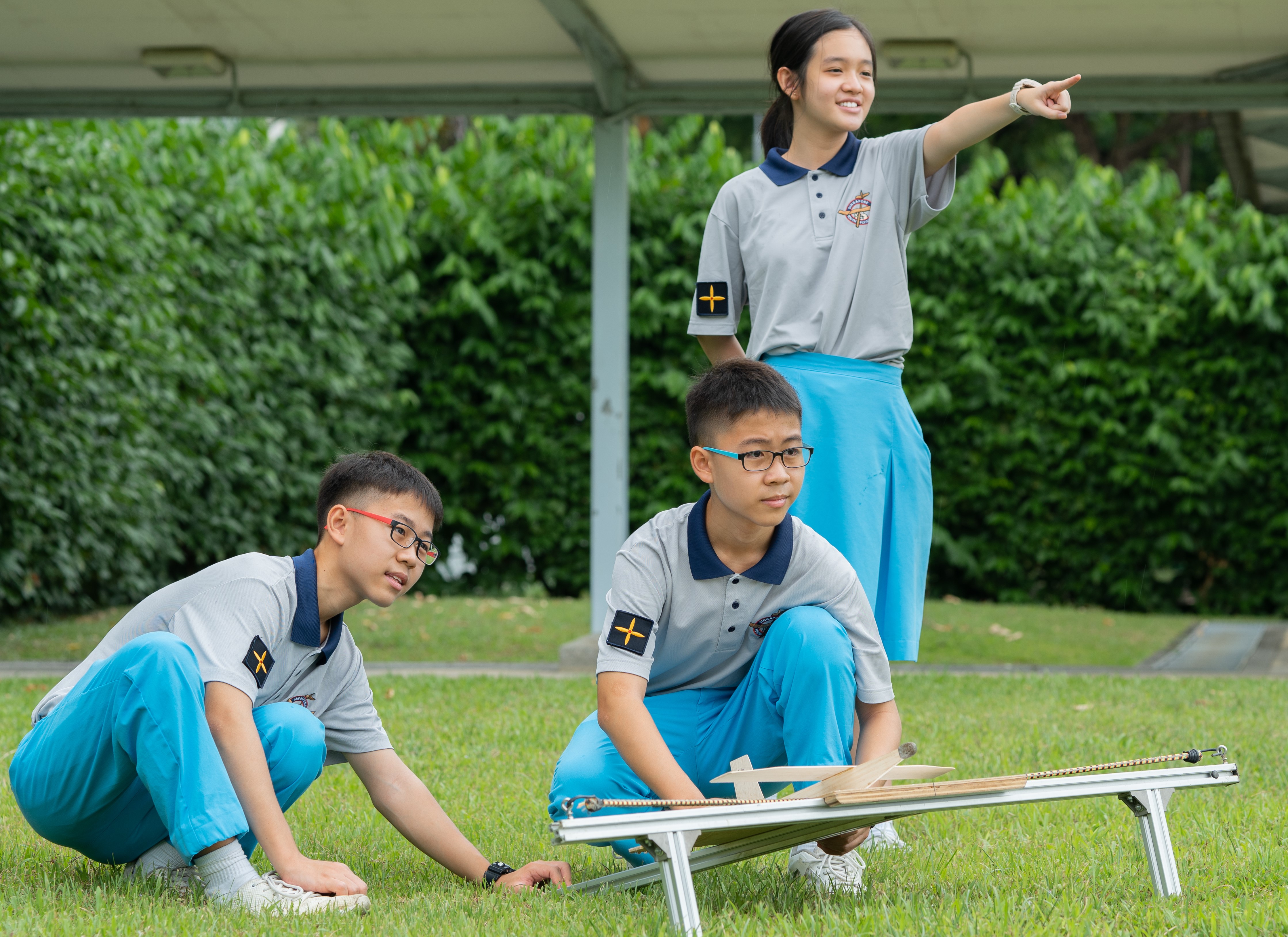Three students in grey and blue uniforms work with a small launcher on grass, one girl pointing ahead.